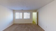 This single-floor residential unit presents a clean, functional layout designed for comfortable everyday living. The living area is spacious and open, finished with neutral-toned wall-to-wall carpeting that extends throughout the main rooms, creating a warm and cohesive feel. Large windows with blinds allow natural light to flow into the space, enhancing the bright, airy atmosphere. A front entry door opens directly into the living area, which also features convenient access to additional rooms via interior doorways.

The galley-style kitchen is efficiently organized with white cabinetry lining both sides, offering ample storage above and below the counter. It is equipped with a white electric range, a refrigerator, a built-in dishwasher, and a double-basin sink, all set on light tile flooring. A ventilation hood above the range adds to the kitchen's practical design.

The bedroom is generously sized and features a wide sliding-door closet with a full-width hanging rod, providing substantial storage. Carpeted flooring continues throughout, maintaining a consistent, comfortable aesthetic.

The bathroom includes a vanity with light wood cabinetry, a white countertop, and a mirror above, complemented by tile flooring. A utility panel is also visible within the space, adding to the unit's self-contained nature. Overall, the layout is practical, well-lit, and move-in ready.