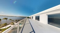 This single-level property is defined by its striking rooftop terrace, which serves as a standout architectural feature. The terrace runs the length of the building and is finished with smooth, light-colored concrete flooring that reflects the abundant natural sunlight. A continuous glass and metal railing system lines the outer edge, providing unobstructed panoramic views of the surrounding waterway. Palm trees dot the shoreline below, reinforcing the tropical coastal setting. The clean white exterior walls of the building face the terrace directly, punctuated by multiple large sliding glass doors that facilitate seamless indoor-outdoor access and allow natural light to flow into the interior spaces. The open-air terrace offers an expansive sense of space and is well-suited for outdoor use throughout the day. The interior spaces, while not fully visible in available imagery, appear to include a bathroom and additional rooms accessible from the terrace level. The overall design emphasizes a modern, minimalist aesthetic with a strong emphasis on outdoor living and the natural waterfront environment that surrounds the property.