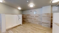 This single-level bathroom is designed with a clean, contemporary aesthetic that prioritizes both functionality and visual appeal. The flooring features wood-look tile planks laid in a linear pattern, lending warmth and a natural character to the space while remaining practical and easy to maintain.

The walls are clad in stacked, multi-toned stone-effect tiles arranged in a horizontal pattern, creating a layered, textural look that adds depth and sophistication. The tile treatment extends across a significant portion of the walls, transitioning to a neutral painted upper section that keeps the overall palette light and airy.

A generously sized open walk-in shower occupies one end of the room, equipped with a handheld showerhead and a built-in bench, making it both accessible and convenient. The open design eliminates the need for a door or curtain, contributing to the spacious feel of the room.

Two flush-mount ceiling light fixtures provide ample, evenly distributed illumination throughout the space. A small window positioned high on the wall allows for a touch of natural light and ventilation. The combination of neutral tones, durable materials, and thoughtful layout makes this bathroom both stylish and highly functional.