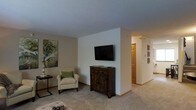 This two-floor space presents a clean, functional design with a welcoming living area as its centerpiece. The living room is finished with neutral-toned carpet flooring and light-colored walls that create a bright, open atmosphere. A wall-mounted flat-screen television is positioned above a rustic dark wood cabinet, which serves as a grounding focal point in the room. A comfortable armchair with a green accent pillow adds a touch of warmth alongside tasteful wall art, including a botanical print that brings a subtle natural element to the space. The textured ceiling adds a classic residential finish throughout. A hallway visible from the living area leads further into the home, lined with framed artwork and providing access to additional rooms, hinting at a well-organized floor plan. Warm wood-toned interior doors complement the overall neutral palette. The layout across two floors offers separation between living and other functional areas, providing versatility for various living arrangements. Overall, the space communicates a relaxed, understated aesthetic with practical design choices suited to everyday living.