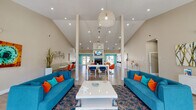 This single-floor community clubhouse makes a striking first impression with its dramatic vaulted ceilings and an open, airy layout filled with natural light. The main lounge area is anchored by two bold teal sofas accented with orange and turquoise throw pillows, arranged around a sleek white coffee table atop a decorative area rug. Tall structural columns frame the space elegantly, adding architectural character throughout.

The open floor plan flows seamlessly from the lounge into a dining and social area, where navy and orange accent chairs surround white high-top and standard tables. A central fireplace with a mounted flat-screen television serves as a focal point, backed by a teal accent wall and large windows that invite views of the outdoors. Wood-look tile flooring runs throughout, tying each zone together cohesively.

The kitchen features light wood cabinetry, white quartz countertops, stainless steel appliances, and globe pendant lighting — all oriented toward a generous window with outdoor views. Adjacent to the main areas, a dedicated business center offers multiple computer workstations along a continuous desk, set against vivid teal walls with stylish globe pendant lights and decorative lattice dividers. The overall ambiance is energetic yet welcoming, with a coastal-inspired color palette that gives the entire space a lively, resort-like character.