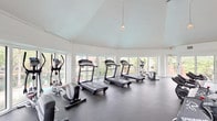 This fitness center is housed in a bright, airy space defined by its striking vaulted ceilings and a sweeping curved wall of large windows that flood the room with natural light and offer open views of the surrounding greenery. The high, tent-like ceiling creates an expansive, open atmosphere, further enhanced by pendant light fixtures that add a clean, functional aesthetic throughout.

The main exercise area is generously proportioned, providing ample space for a well-rounded selection of cardio equipment, including multiple treadmills, elliptical trainers, and spinning bikes. A dedicated strength training zone features a multi-function cable machine and weight bench, catering to a full range of workout routines.

The interior is finished with durable dark rubber flooring, providing both safety and practicality for high-impact workouts. Crisp white walls and trim throughout contribute to the clean, modern feel of the space. A built-in shelving and cabinetry unit along one wall provides convenient storage, while a wall-mounted television adds an entertainment option for users. A private entry door and visible ventilation features ensure comfort and accessibility. Overall, the fitness center combines functional design with an inviting, light-filled environment ideal for daily exercise.