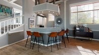 Located at 100 Oakwood Dr in Bethel Park, PA, this thoughtfully designed single-floor community space blends contemporary style with comfortable, functional living. The main lounge area serves as the centerpiece, boasting soaring coffered ceilings with dark-painted recessed panels, recessed lighting, and a ceiling fan that enhance the room's impressive vertical scale. A floor-to-ceiling stone fireplace anchors the space, topped with a rustic wood mantel and a wall-mounted TV, while decorative circular wall art adds an artistic focal point. Plush seating, including teal velvet accent chairs and leather sofas, surrounds a bold geometric-patterned area rug, creating an inviting gathering atmosphere. Adjacent to the lounge, a bar-style kitchen area features a large granite-topped island with leather barstool seating, white cabinetry, and ample counter space—ideal for casual socializing. Wide-plank hardwood-style flooring flows consistently throughout, unifying each zone. A game area with a shuffleboard table and glazed interior partitions adds recreational variety, while large windows dressed with wood blinds flood the entire space with generous natural light and views of the surrounding outdoors.