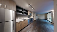 Located in Seattle's vibrant Capitol Hill neighborhood, Unit 316 at 721 E Pine Street offers a thoughtfully designed, single-floor residence with a contemporary aesthetic throughout. The open-concept kitchen and living area serves as the heart of the unit, featuring rich dark hardwood-style flooring and a well-appointed kitchen equipped with stainless steel appliances — including a full-size refrigerator, electric range, dishwasher, and over-the-range microwave. Warm wood-toned cabinetry is complemented by sleek white quartz countertops and open upper shelving, lending a modern, urban feel. Track lighting illuminates the kitchen and entry area efficiently. The living space benefits from large floor-to-ceiling windows and a sliding glass door that flood the room with natural light and open onto views of a lush, landscaped courtyard, creating a calm and inviting atmosphere. The bathroom is finished with the same modern sensibility, featuring a wide floating vanity with ample storage, clean tile flooring, and a glass-enclosed shower. Wall sconces provide warm, accent lighting. Throughout the unit, neutral tones, clean lines, and quality finishes create a cohesive and functional living environment well-suited to urban life in Seattle.