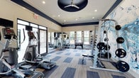 This modern fitness center is thoughtfully designed to accommodate a full range of workout routines. The space features a stylish interior with a bold navy and cream color scheme accented by crown molding along the upper walls. Recessed lighting and a striking circular ceiling fan provide both illumination and air circulation throughout the room. Large windows along the perimeter flood the space with natural light and offer views of the surrounding greenery, creating an energizing atmosphere for exercise. Floor-to-ceiling mirrors line one wall, enhancing the sense of openness and allowing users to monitor their form. The flooring consists of durable, multi-toned carpet tiles in blue and grey hues, providing both comfort and functionality. Equipment is well-organized and includes cardio machines such as ellipticals and rowing machines, as well as a comprehensive strength training station with cable attachments and weight plates. A dedicated free weights area features a full rack of dumbbells alongside an adjustable bench. The space is completed with a mounted flat-screen television, making it a fully functional and comfortable fitness facility suitable for a wide variety of exercise needs.