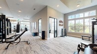 This expansive fitness center is thoughtfully designed to accommodate a wide range of workout styles and fitness goals. The main exercise floor is generously proportioned, featuring plush patterned carpeting and soaring ceilings with recessed lighting that create an open, energizing atmosphere. Large windows line the walls, flooding the space with natural light and offering pleasant outdoor views. The main floor houses an impressive array of cardio equipment, including ellipticals, stair climbers, and rowing machines, alongside a dedicated strength training area equipped with cable machines, weight benches, a full dumbbell rack, and a punching bag. A striking textured accent wall adds visual depth to the strength zone, which is further enhanced by floor-to-ceiling mirrors. A separate studio room, accessed through sleek glass-paneled doors with black frames, features light hardwood flooring, a wall-mounted television, and storage for stability balls, resistance equipment, and accessories — ideal for group classes or personal training sessions. The studio also benefits from tall transom windows that amplify the sense of airiness. Throughout, the neutral warm-toned palette, clean architectural lines, and high-end finishes combine to create a modern, welcoming environment suited for comprehensive fitness activities.