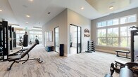 This expansive fitness center is thoughtfully designed to accommodate a wide range of workout styles and fitness goals. The main exercise floor is generously proportioned, featuring plush patterned carpeting and soaring ceilings with recessed lighting that create an open, energizing atmosphere. Large windows line the walls, flooding the space with natural light and offering pleasant outdoor views. The main floor houses an impressive array of cardio equipment, including ellipticals, stair climbers, and rowing machines, alongside a dedicated strength training area equipped with cable machines, weight benches, a full dumbbell rack, and a punching bag. A striking textured accent wall adds visual depth to the strength zone, which is further enhanced by floor-to-ceiling mirrors. A separate studio room, accessed through sleek glass-paneled doors with black frames, features light hardwood flooring, a wall-mounted television, and storage for stability balls, resistance equipment, and accessories — ideal for group classes or personal training sessions. The studio also benefits from tall transom windows that amplify the sense of airiness. Throughout, the neutral warm-toned palette, clean architectural lines, and high-end finishes combine to create a modern, welcoming environment suited for comprehensive fitness activities.