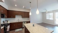 This single-floor residence showcases a thoughtfully designed open-concept layout connecting the kitchen, dining, and living areas. The kitchen is a standout feature, equipped with rich dark-walnut cabinetry, light granite countertops, and a generous central island illuminated by elegant teardrop pendant lights. Stainless steel appliances — including a gas range, built-in microwave, French door refrigerator, and dishwasher — provide a fully functional cooking space. Wood-look plank flooring runs throughout the main living areas, adding warmth and continuity. A stacked washer and dryer unit is conveniently integrated near the kitchen. The living area benefits from natural light through large windows and a glass-paneled exterior door, complemented by a ceiling fan overhead. The bedroom offers plush carpeting, dual windows with blinds, and its own ceiling fan, creating a comfortable, well-lit retreat. The bathroom features a combined tub and shower with a curved rod, built-in shelving, wood-look flooring, and clean neutral finishes. Throughout the unit, neutral wall tones and recessed lighting contribute to a bright, cohesive, and contemporary ambiance.