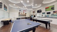 Located at 2015 Wave Crest Dr in Raleigh, NC, this single-floor entertainment space is purpose-built for recreation and social gatherings. The room immediately draws the eye upward to its dramatic coffered ceiling, composed of layered octagonal panels accented with recessed lighting and crisp crown molding — a refined architectural detail that elevates the overall aesthetic.

Rich, dark hardwood flooring runs throughout, providing a warm contrast to the light-toned walls. The space is generously proportioned to accommodate an impressive array of gaming options: a full-sized pool table with cue rack, a regulation ping-pong table, a foosball table, a shuffleboard table, and multiple classic arcade and pinball machines. Multiple wall-mounted flat-screen televisions are positioned for easy viewing from nearly any area of the room.

A built-in cabinetry and countertop section with dark wood finish serves as a bar and service area, complemented by bar-height seating for casual socializing. Large windows along one wall invite natural light and offer views of the surrounding greenery. Full-length mirrors along another wall amplify the sense of space and light. Vibrant billiards-themed artwork decorates the walls, reinforcing the room's energetic, game-focused character. This space is thoughtfully designed for maximum entertainment versatility.