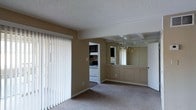 This single-floor residence offers a comfortable and functional layout with a cohesive, neutral aesthetic throughout. The main living and dining area is carpeted and bathed in natural light through large vertical-blind-covered sliding glass doors, creating a bright and open atmosphere. A textured ceiling adds depth to the space, while exposed decorative beams lend a subtle architectural charm.

The dining area is distinguished by a full-wall mirrored panel that visually expands the room and reflects a brass chandelier, adding a classic, elegant touch. A wainscoted half-wall separates and defines this space without closing it off, maintaining an open, connected feel.

An open doorway leads directly into the kitchen, which features dark upper cabinetry contrasted by a white range and classic appliances. A stone tile backsplash adds texture and visual interest to the cooking area. The warm, tan-toned walls carry consistently through the space, tying the rooms together with a unified, inviting color palette. Overall, the layout is practical and well-defined, offering distinct living, dining, and kitchen zones within a single-level footprint.