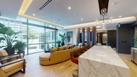 Located at 419 SE Second St in Fort Lauderdale, FL, this upscale single-floor space exemplifies contemporary design with a refined, hospitality-inspired aesthetic. The main lounge area is anchored by a striking white marble-veined counter, complemented by warm light wood flooring that runs throughout. Tall floor-to-ceiling glass walls flood the space with abundant natural light while offering views of lush palm trees and the outdoor surroundings, creating a seamless indoor-outdoor connection.

Richly toned vertical wood panel dividers elegantly partition the open layout into distinct seating zones, each thoughtfully furnished with designer pieces — including curved sofas, velvet accent chairs in deep blue, and sculptural occasional tables. Recessed cove lighting and globe pendant chandeliers add a warm, layered glow to the tray ceilings above.

A quieter lounge alcove features charcoal walls contrasted by light furnishings, sculptural art pieces, and framed geometric wall art, lending an intimate gallery-like quality. Elsewhere, a curated wall display of bold black-and-white abstract canvases anchors a casual seating nook with low-profile chairs and metallic accent stools. A serene windowed nook with sheer draping provides a private retreat. Lush tropical plants are thoughtfully placed throughout, reinforcing the vibrant South Florida ambiance.