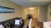 This single-floor space offers a well-designed open-concept layout that seamlessly connects the living and dining areas, creating a cohesive and functional environment. The living area is anchored by a comfortable dark grey sofa accented with decorative pillows, complemented by a patterned area rug that adds texture to the hardwood-style flooring running throughout the space. Warm, neutral beige walls provide a welcoming backdrop and contribute to the overall sense of warmth and continuity.

The dining area is positioned toward the rear of the open space and features a round dark wood table surrounded by matching cross-back chairs, ideal for intimate gatherings. Overhead, a modern track lighting fixture illuminates the dining zone effectively, while playful wall-mounted utensil décor adds a charming, culinary-themed touch to the area. A framed botanical wall art piece near the living area contributes an additional layer of visual interest.

White baseboards provide a clean contrast against the flooring, and the smooth ceiling with subtle texture adds to the polished finish of the space. Overall, the layout is practical, modern, and thoughtfully styled with a consistent neutral palette.
