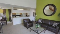 This two-floor space is designed with an open-concept layout that maximizes the sense of volume and airiness throughout. The living area is anchored by dramatically high ceilings that draw the eye upward, creating an expansive atmosphere. A striking lime-green accent wall adds a bold pop of color and serves as a focal point, complemented by a decorative round mirror that enhances the visual depth of the room. The living space is carpeted and comfortably accommodates seating, including a dark leather sofa and matching armchair arranged around a glass-topped coffee table, creating an inviting and functional gathering area.

The kitchen is seamlessly connected to the living space via a pass-through counter with a breakfast bar, allowing for easy interaction between the two areas. The kitchen is equipped with standard appliances, including a refrigerator and microwave, and features wood-toned cabinetry with ample storage. Overhead lighting illuminates the kitchen efficiently. The open sightlines between the kitchen and living room contribute to the cohesive, connected feel of the main floor, making the overall layout practical and well-suited for everyday living and entertaining.