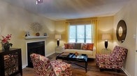 Located at 8000 Waters Ave in Savannah, Georgia, this single-floor residence offers a thoughtfully designed living environment with a welcoming and cohesive aesthetic. The living area serves as the heart of the home, showcasing warm golden-yellow walls that complement rich hardwood-style flooring. A classic white fireplace with a decorative mantel anchors the room, adding both a functional focal point and timeless charm. Natural light pours in through a large window dressed with bold orange-toned curtains, creating a bright and energizing atmosphere. The space is well-suited for both relaxed living and entertaining, with ample room for comfortable seating arrangements. Warm earth tones and patterned accents carry throughout, contributing to a cohesive and vibrant interior palette. The single-floor layout offers convenient, accessible living with a practical flow between spaces. Situated in the established Savannah, GA community near Waters Avenue, this residence benefits from a well-connected location with access to local amenities, dining, and the rich cultural heritage of the surrounding area. Overall, the space combines functional design with a warm, inviting ambiance that reflects the character of its Savannah setting.