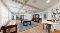 Located at 13220 North Scottsdale Road in Scottsdale, Arizona, this thoughtfully designed amenity space combines upscale entertainment and functional gathering areas within a single floor. The game room serves as the centerpiece, showcasing a full-size pool table anchored on a vibrant teal area rug, a shuffleboard table, and a café-style high-top seating area. An elegant coffered ceiling with recessed lighting and a geometric pendant chandelier adds architectural distinction, while warm terracotta-toned brick flooring lends a welcoming, textured ambiance. Large windows invite natural light, and glass-paneled French doors provide access to adjacent private meeting or lounge rooms, visible through the partitions.

The kitchen is a standout feature, designed with rich dark wood cabinetry, an intricate patterned tile backsplash, and a premium white range beneath a cream-and-gold range hood. Exposed wooden ceiling beams add rustic character, while a generously sized island topped with a striking green marble-style counter provides ample workspace and bar seating. A multi-arm chandelier illuminates the kitchen with warm, layered light, creating an inviting space well-suited for catered events or community gatherings. Together, these spaces form a cohesive, upscale environment built for both recreation and connection.