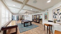 Located at 13220 North Scottsdale Road in Scottsdale, Arizona, this thoughtfully designed amenity space combines upscale entertainment and functional gathering areas within a single floor. The game room serves as the centerpiece, showcasing a full-size pool table anchored on a vibrant teal area rug, a shuffleboard table, and a café-style high-top seating area. An elegant coffered ceiling with recessed lighting and a geometric pendant chandelier adds architectural distinction, while warm terracotta-toned brick flooring lends a welcoming, textured ambiance. Large windows invite natural light, and glass-paneled French doors provide access to adjacent private meeting or lounge rooms, visible through the partitions.

The kitchen is a standout feature, designed with rich dark wood cabinetry, an intricate patterned tile backsplash, and a premium white range beneath a cream-and-gold range hood. Exposed wooden ceiling beams add rustic character, while a generously sized island topped with a striking green marble-style counter provides ample workspace and bar seating. A multi-arm chandelier illuminates the kitchen with warm, layered light, creating an inviting space well-suited for catered events or community gatherings. Together, these spaces form a cohesive, upscale environment built for both recreation and connection.
