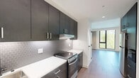 Located at 31 Artful Way in Asheville, NC, this thoughtfully designed single-floor unit offers a compact yet efficient layout encompassing a living room, kitchen, bathroom, hallway, and two closets. The kitchen is a standout feature, showcasing dark espresso cabinetry that extends to the ceiling, crisp white quartz countertops, stainless steel appliances including an electric range and refrigerator, and a striking hexagonal tile backsplash. Recessed lighting and wood-look flooring run throughout, lending a cohesive, modern aesthetic to the entire space. The open living area is bathed in natural light, courtesy of large black-framed floor-to-ceiling windows that frame lush tree canopy views, creating a bright and airy atmosphere. Neutral-toned drapes add softness to the clean-lined interior. The bathroom is finished in a minimalist style with large-format white tile flooring, a floating vanity with a concrete-look basin set on a matte black frame, subway-style wall tile, and a glass-enclosed shower. Two closets provide ample storage. The overall design balances modern sophistication with functional efficiency, making excellent use of every square foot in this well-appointed Asheville residence.