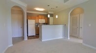 This single-floor residence showcases an open-concept layout that seamlessly connects the kitchen and living areas. The kitchen is equipped with warm honey-toned cabinetry, white appliances including a full-size refrigerator and built-in microwave, and a dark-toned range. A peninsula with a stone-finish countertop serves as both a workspace and a natural divider between the kitchen and the adjoining living space, which features neutral-toned carpet flooring. The kitchen itself has hardwood-style flooring, creating a practical and visually distinct transition between zones. Two pendant lights hang above the peninsula, adding focused task lighting and a decorative touch. Crown molding runs along the upper perimeter of the space, lending a refined, polished character to the interiors. Elegant arched doorways frame the entrances on either side of the living area, contributing architectural interest and an open, airy feel. The neutral color palette of warm beige tones on the walls complements the overall design, creating a cohesive and welcoming atmosphere throughout the space.