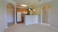This single-floor residence showcases an open-concept layout that seamlessly connects the kitchen and living areas. The kitchen is equipped with warm honey-toned cabinetry, white appliances including a full-size refrigerator and built-in microwave, and a dark-toned range. A peninsula with a stone-finish countertop serves as both a workspace and a natural divider between the kitchen and the adjoining living space, which features neutral-toned carpet flooring. The kitchen itself has hardwood-style flooring, creating a practical and visually distinct transition between zones. Two pendant lights hang above the peninsula, adding focused task lighting and a decorative touch. Crown molding runs along the upper perimeter of the space, lending a refined, polished character to the interiors. Elegant arched doorways frame the entrances on either side of the living area, contributing architectural interest and an open, airy feel. The neutral color palette of warm beige tones on the walls complements the overall design, creating a cohesive and welcoming atmosphere throughout the space.