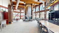 Located at 20200 Poplar Creek Pkwy in Brookfield, Wisconsin, this generously sized single-floor living space serves as an impressive communal gathering area. The open-concept layout is anchored by dramatic soaring ceilings with exposed wooden beam framework, lending a warm yet grand architectural character throughout.

Floor-to-ceiling windows line the perimeter, flooding the interior with abundant natural light and offering expansive views of the surrounding outdoor patio and courtyard, complete with umbrella-shaded seating. The large window walls are framed by rich, dark wood structural elements that add depth and visual interest.

A standout feature is the striking double-sided stone and tile fireplace column at the heart of the space, creating a cozy focal point surrounded by contemporary white lounge chairs and a round side table. Adjacent seating areas include a large sectional sofa and accent chairs positioned to take full advantage of the outdoor views.

The space also includes a full bar and counter area featuring dark cabinetry, open shelving, and bar stool seating set against bold red accent walls, adding a vibrant, energetic contrast to the otherwise neutral palette. A separate dining nook with red leather chairs and café-style tables offers an intimate setting near the windows.

Soft, textured carpeting throughout ties all the zones together, creating a cohesive and inviting atmosphere well-suited for both social gatherings and relaxed individual use.