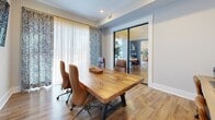 This single-floor space is designed as a functional yet stylish meeting or private office environment. The room centers around a substantial solid wood conference table with a live-edge character finish, complemented by ergonomic leather rolling chairs that seat multiple occupants comfortably. Warm-toned hardwood flooring runs throughout, contributing to an inviting, professional atmosphere.

Natural light filters generously into the space through large windows dressed with layered treatments—sheer white panels paired with bold, patterned drapery in navy and white, adding a distinctive decorative touch. A wall-mounted flat-screen display is positioned for easy viewing from the table, supporting presentations and collaborative work.

A sleek glass-paneled door with a black metal frame serves as the primary entry, offering visual transparency while maintaining a sense of separation from adjacent areas. The door connects to a short transitional hallway, flanked by clean white walls and bright baseboards, reinforcing the space's crisp, contemporary aesthetic.

Recessed lighting and a ceiling-mounted HVAC vent ensure practical comfort, while the curved wall detail adds subtle architectural interest. The overall design balances functionality with refined, modern style.