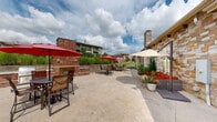 Located at 12302 S Yukon Ave in Glenpool, Oklahoma, this generously sized outdoor patio area serves as a well-appointed communal gathering space. The stamped concrete surface spans a broad, open area that accommodates multiple distinct seating zones, creating a versatile environment for both relaxed lounging and casual dining.

A standout architectural feature is the striking brick outdoor fireplace, which anchors one section of the patio and adds a warm, social focal point. Nearby, a built-in grill station provides a convenient outdoor cooking setup. Vibrant red market umbrellas are positioned throughout the patio, offering shade across bar-height bistro tables and chairs, while a neutral-toned umbrella shelters a comfortable outdoor lounge set with bold red cushions.

The perimeter of the patio is lined with lush, mature landscaping, including tall columnar evergreens and a variety of tropical and ornamental plants that provide both privacy and greenery. An iron fence separates the patio from an adjacent pool area, which is visible in the background alongside manicured lawn space.

The adjacent building showcases attractive stone and siding exterior construction with a pitched shingle roof and a prominent stone chimney. The overall ambiance is inviting and resort-like, designed to encourage outdoor living and community interaction under the open Oklahoma sky.