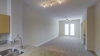 This single-story space centers around a comfortable and thoughtfully arranged living area that exudes a calm, neutral aesthetic. The room benefits from notably high ceilings, contributing to an open and airy atmosphere that enhances the overall sense of space. A ceiling fan with an integrated light fixture is mounted overhead, providing both practical air circulation and subtle illumination.

Natural light is a key feature, streaming in generously through a large glass door dressed with sheer, light-filtering curtains that soften the brightness while maintaining an inviting glow. The room is carpeted in a neutral tone that complements the warm beige walls, creating a cohesive and understated palette throughout.

The living area is furnished with a tufted sofa in a soft taupe, accompanied by a dark upholstered armchair, wicker accent chairs, and a square ottoman that doubles as a coffee table. Ambient lighting is provided by matching table lamps positioned on either side of the seating arrangement. Decorative touches, including framed wall art and a tall botanical accent piece, add visual interest without overwhelming the space. Overall, the room presents a clean, functional, and welcoming environment.