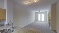 This single-story space centers around a comfortable and thoughtfully arranged living area that exudes a calm, neutral aesthetic. The room benefits from notably high ceilings, contributing to an open and airy atmosphere that enhances the overall sense of space. A ceiling fan with an integrated light fixture is mounted overhead, providing both practical air circulation and subtle illumination.

Natural light is a key feature, streaming in generously through a large glass door dressed with sheer, light-filtering curtains that soften the brightness while maintaining an inviting glow. The room is carpeted in a neutral tone that complements the warm beige walls, creating a cohesive and understated palette throughout.

The living area is furnished with a tufted sofa in a soft taupe, accompanied by a dark upholstered armchair, wicker accent chairs, and a square ottoman that doubles as a coffee table. Ambient lighting is provided by matching table lamps positioned on either side of the seating arrangement. Decorative touches, including framed wall art and a tall botanical accent piece, add visual interest without overwhelming the space. Overall, the room presents a clean, functional, and welcoming environment.