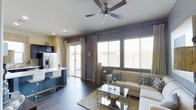 This single-story space showcases an open-concept layout that seamlessly connects the kitchen and living areas, creating a cohesive and functional environment. The living room is a focal point, anchored by large windows fitted with white plantation shutters that allow generous natural light to fill the room. A sliding glass door opens to an outdoor area, further enhancing the bright, airy feel. The walls feature a sophisticated two-tone color scheme, with cool slate-blue accents contrasting against crisp white surfaces.

The living area is designed for comfort and style, centered around a cream-toned sectional sofa adorned with patterned throw pillows. A glass-top coffee table and a coordinating etagere add an elegant, contemporary touch, while a plush area rug grounds the seating arrangement. A ceiling fan with dark wooden blades is mounted overhead, adding both function and visual interest.

The kitchen is efficiently designed, featuring a stainless steel refrigerator with an ice and water dispenser, dark upper cabinetry, and a blue kitchen island with a white countertop accompanied by modern bar seating. Dark hardwood-style flooring runs throughout, tying the entire space together cohesively.