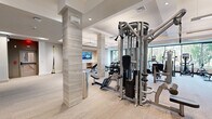 Located at 99 Wilkens Lane in Barnstable, MA, this fitness center offers a generously sized, single-floor layout designed to accommodate a comprehensive range of workout needs. The space features a modern, upscale aesthetic defined by coffered ceilings with recessed and pendant lighting, textured stone-clad columns, and neutral-toned carpeting that lends a polished yet functional feel throughout.

The facility is thoughtfully organized into distinct workout zones. A dedicated cardio area lines one side of the room with multiple treadmills, ellipticals, and stair climbers, positioned near large windows that allow abundant natural light and views of the surrounding greenery. A free weights section features a full rack of dumbbells alongside adjustable benches, ideal for strength training. Multi-use cable machines and a leg press station provide a complete strength circuit, while a TRX suspension training station supports functional fitness routines.

Floor-to-ceiling mirrors along one wall visually expand the space and assist with form and technique. A wall-mounted television near a built-in cabinetry unit adds an entertainment element to the environment. The clean, contemporary design, combined with ample floor space between equipment, ensures a comfortable and professional workout experience suited for a wide range of fitness levels.
