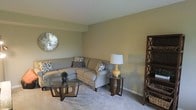 This two-floor space presents a well-arranged living area characterized by soft, neutral wall tones and wall-to-wall carpeting that contribute to a calm and comfortable ambiance. The living room is anchored by a generously sized sectional sofa in a warm beige upholstery, accessorized with patterned throw pillows that introduce subtle visual interest. A glass-topped coffee table with a distinctive dark wood base serves as a functional centerpiece, while a matching side table holds a ceramic table lamp with a warm-toned base, providing soft ambient lighting. Open shelving in a dark wood finish stands along one wall, offering display and storage space. A decorative metallic sunburst mirror adorns the wall, adding a reflective accent that enhances the sense of space. The carpeted flooring runs throughout, lending a soft underfoot feel to the room. The overall design aesthetic leans toward transitional style, blending classic furnishing silhouettes with contemporary decorative touches. The layout suggests a practical and livable space, with the second floor likely offering additional private living areas.