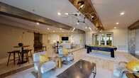 Located at 900 Hobbs Road in Greensboro, NC, this single-floor community clubhouse is a thoughtfully designed social hub that blends modern elegance with comfortable gathering spaces. The open-concept main lounge serves as the centerpiece, featuring light wood-style tile flooring, a striking sculptural chandelier with interlocking rings, and an expansive layout that accommodates multiple seating arrangements. A dedicated lounge area anchors one end of the space, where a sleek wall-mounted flat-screen TV sits above a linear gas fireplace framed by a warm, backlit tile surround — ideal for relaxed gatherings. Adjacent to the main lounge, a fully equipped bar area stands out with a large marble-topped island, dark wood cabinetry, globe pendant lighting, a coffee station, and a wall-mounted TV set against a metallic mosaic tile backsplash. A billiards table adds a recreational focal point to the central area, complemented by high-top bistro seating near floor-to-ceiling windows that invite natural light. Tucked away is a cozy niche lounge with exposed brick-style walls, built-in bench seating with plush cushions, pendant lighting, and a chalkboard — offering a more intimate retreat. Throughout, the design harmonizes warm gold accents, neutral tones, and curated décor to create a welcoming, upscale atmosphere.