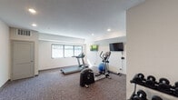 This single-floor fitness center at 175 18th St SW in Owatonna, Minnesota, is a well-equipped exercise space designed to support a well-rounded workout routine. The room features neutral-toned walls, dark base molding, and durable carpet flooring throughout, creating a clean and functional environment. Recessed lighting is evenly distributed across the ceiling, ensuring the space is well-lit at all times, while a row of exterior-facing windows along one wall allows natural light to filter in and provides views of the surrounding outdoor area.

The fitness center is outfitted with a variety of cardio equipment, including a Cybex treadmill and an elliptical cross-trainer, positioned to take advantage of the natural light. A multi-tiered dumbbell rack stocked with a full range of free weights occupies one corner, offering ample options for strength training. An adjustable workout bench provides a dedicated space for resistance exercises. A stability ball rounds out the functional fitness accessories available. Multiple flat-screen televisions are mounted on the walls, allowing users to stay entertained during their workouts. The layout is thoughtfully arranged to maximize usable floor space, leaving room for stretching or floor exercises.