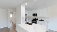This single-floor space showcases a well-appointed kitchen designed with a clean, contemporary aesthetic. The kitchen features white shaker-style cabinetry that extends to the upper walls, providing ample storage while contributing to the light, airy atmosphere throughout. A standout element is the striking quartz peninsula countertop, distinguished by its subtle veining pattern, which houses an undermount stainless steel sink with a modern pull-down faucet. This peninsula creates a natural division between the kitchen and the adjoining hallway area, offering both workspace and a casual gathering point. Stainless steel appliances, including a gas range, over-the-range microwave, and refrigerator, are seamlessly integrated into the cabinetry, adding a polished, professional touch. Recessed lighting illuminates the space evenly, complementing the white walls and light wood-toned flooring that runs throughout. The hallway beyond the kitchen provides a glimpse into additional rooms, suggesting a practical and well-connected layout. Overall, the space presents a bright, functional, and modern environment suited for everyday living.