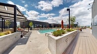 This expansive outdoor amenity space is designed with a contemporary aesthetic, characterized by clean lines, dark-toned architectural elements, and thoughtfully curated landscaping. At the heart of the space is a large, resort-style swimming pool equipped with an accessibility lift, surrounded by an open concrete sundeck lined with modern chaise lounges and vibrant orange accent umbrellas. Adjacent to the main pool, a separate hot tub provides a more intimate relaxation area, flanked by additional lounge seating beneath a series of architectural pergola structures. Raised concrete planters filled with native grasses and ornamental plantings add texture and visual interest throughout the deck. A dedicated turf lawn area serves as a versatile social hub, featuring Adirondack chairs, string lights overhead, gas grills, and a whimsical vintage-style camper accent piece. A large outdoor LED screen anchors one end of the lawn, creating an ideal outdoor cinema or sports-viewing experience. A covered pergola with bar-height seating offers a shaded gathering spot with panoramic mountain views stretching across the horizon. The entire space enjoys abundant natural light and open skies, combining functional amenities with a vibrant, resort-inspired atmosphere.