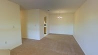 This single-floor space presents an open and functional layout designed for comfortable everyday living. The main living area is finished with wall-to-wall carpeting in a warm neutral tone, complementing the cream-colored walls and white baseboards that run throughout. A modest chandelier hangs centrally, adding a subtle decorative touch to the dining or living zone. The ceiling features a light texture and includes a visible air vent, indicating a standard HVAC system. A partial interior wall creates a natural division within the open floor plan, helping to define separate functional zones without fully closing off the space. Through a doorway, the kitchen is accessible and features wood-toned cabinetry, light-colored flooring, and what appears to be a range or stove, providing a practical cooking area. The overall layout flows efficiently between the living and kitchen spaces. The neutral palette and open arrangement offer a versatile canvas, making the space adaptable for various furnishing and design preferences. The single-floor configuration ensures easy accessibility throughout.