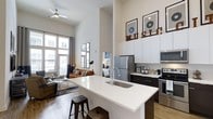 This stylish single-floor unit showcases a thoughtfully designed open-concept layout that seamlessly connects the kitchen, dining, and living areas into one cohesive, airy space. The dramatically high ceilings amplify the sense of volume and grandeur, while large windows flood the interior with generous natural light, offering views of the surrounding urban streetscape.

The contemporary kitchen is a standout feature, equipped with stainless steel appliances including a refrigerator, microwave, and range. Sleek two-tone cabinetry — combining dark lower cabinets with crisp white upper cabinets — provides ample storage, while a spacious white quartz kitchen island with an undermount sink and bar seating serves as both a functional workspace and an inviting gathering spot.

The living area extends naturally from the kitchen, anchored by warm hardwood-style flooring that runs throughout the open space. A ceiling fan enhances comfort, and the overall décor reflects a modern urban aesthetic, complete with music-themed artwork accents that add personality and character to the space. The cohesive design, elevated ceilings, and abundant light combine to create a bright, welcoming atmosphere ideal for both everyday living and entertaining.