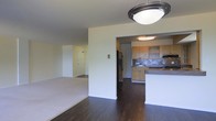 This single-floor residence offers a practical and well-organized open-concept layout that connects the main living spaces with ease. The main living area features rich, dark hardwood flooring that extends throughout the common spaces, creating a warm and cohesive aesthetic. A ceiling-mounted light fixture anchors the central area, providing ambient illumination across the open floor plan. Adjacent to the living space, a carpeted room offers a softer, more comfortable environment, suitable for use as a bedroom or secondary lounge area. The kitchen is a functional highlight of the home, outfitted with light wood cabinetry, a mosaic tile backsplash, dark granite-style countertops, and stainless steel appliances including a refrigerator and built-in microwave. A peninsula countertop extends into the open area, offering additional workspace and serving as a natural divider between the kitchen and the rest of the living space. The kitchen is finished with its own ceiling light, ensuring a bright workspace. Throughout the residence, neutral wall tones keep the interior feeling clean and versatile, providing a ready canvas for personalization. The overall layout is efficient, making excellent use of the available space across a single level.