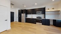 This contemporary single-floor unit showcases a thoughtfully designed open-concept layout where the kitchen and living area flow seamlessly together. Light hardwood floors extend throughout, providing a warm contrast to the modern black-framed windows and doors. The kitchen is a standout feature, equipped with floor-to-ceiling matte black cabinetry accented by gold hardware, a white subway tile backsplash, quartz countertops, a gold-finish faucet, and a full suite of stainless steel appliances. Open shelving adds a functional, airy touch to the design. The spacious living area is bathed in natural light from multiple black-framed windows and sliding glass doors that open onto a private balcony with urban views. A sleek ceiling fan with a matte black finish adds both style and comfort to the living space. A sliding barn door provides a striking architectural detail that separates spaces with character. The bathroom is elegantly finished with dark hexagonal tile flooring, a double-sink vanity featuring matte black fixtures and gold-accented hardware, a tub-shower combination enclosed by a bold black grid-frame glass door, and backlit mirrors. The bedrooms continue the clean, minimalist aesthetic with hardwood floors, black-framed windows, and ceiling fans.
