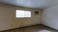 This single-floor space features a simply designed room finished with neutral, cream-colored walls that create a clean and understated atmosphere. The room is covered in wall-to-wall carpet in a muted, earthy tone, providing a practical and comfortable underfoot surface. A horizontal window fitted with white horizontal blinds allows natural light to filter into the space, offering a view of the outdoors. The textured popcorn ceiling adds a classic touch characteristic of older residential construction. Climate control is well-addressed, with both a baseboard heating unit running along the exterior wall beneath the window and a wall-mounted air conditioning unit positioned nearby, ensuring year-round comfort. The overall layout is open and unfurnished, presenting a blank canvas that can accommodate a variety of uses. The room's straightforward design emphasizes functionality, making it a versatile space suited to a range of configurations.