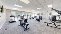 Located at 3901 Noyes Circle in Randallstown, Maryland, this fitness center offers a generously sized, single-floor exercise room designed to support a full range of workout routines. The space features a clean, modern aesthetic with neutral gray walls, a suspended tile ceiling with recessed panel lighting, and durable patterned carpet flooring throughout.

Natural light flows in abundantly through multiple windows fitted with white shutters and blinds, as well as large sliding glass doors that open to an outdoor view — creating a bright and energizing atmosphere. A wall-mounted flat-screen television adds a multimedia element to the cardio area.

The facility is well-equipped with a diverse array of professional-grade machines. The cardio section includes multiple treadmills, upright and recumbent stationary bikes, elliptical trainers, and a spinning bike. The strength training area features Life Fitness cable and resistance machines, a multi-function weight station, and a rowing machine. Fitness accessories such as yoga/exercise mats are also available. Motivational fitness posters and a water dispenser round out the amenities.

The layout provides ample open floor space between equipment, ensuring comfortable movement throughout the room. The overall ambiance is clean, functional, and welcoming — well-suited for daily fitness use.