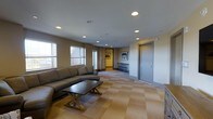 The lounge at 205 Church St in New Haven, Connecticut, is a well-appointed, single-floor gathering space designed for comfort and functionality. The room features a neutral color palette of warm beige and gray tones, creating a calm and inviting atmosphere. Recessed lighting is distributed evenly across the smooth ceiling, providing consistent, well-lit illumination throughout the space.

A large sectional sofa in a charcoal gray upholstery serves as the centerpiece of the seating area, paired with a sturdy wooden coffee table that anchors the arrangement. The floor is covered in a patterned carpet with a subtle checkerboard design in complementary tan and gray hues, adding texture and visual interest underfoot.

Multiple windows along one wall allow natural light to filter into the space, offering views of the surrounding exterior. Framed artwork adorns the walls, contributing a refined aesthetic to the room. A hallway connects to additional areas, suggesting the lounge is part of a larger facility. The overall design balances practicality with a polished, comfortable ambiance suited for communal use.
