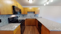 This second-floor 2-bedroom, 1-bathroom unit offers a thoughtfully designed open-concept layout that seamlessly connects the living area to the kitchen. The main living space features rich hardwood-style flooring that extends throughout, creating a warm and cohesive feel. A peninsula counter with granite countertops serves as a natural divider between the kitchen and living area, providing additional workspace and casual seating potential.

The kitchen is well-equipped with warm honey-toned wood cabinetry, granite countertops, a decorative tile backsplash, and black appliances including a range, over-the-range microwave, dishwasher, and refrigerator. Track and flush-mount lighting illuminate the space effectively.

Both bedrooms are carpeted and feature large windows that allow natural light and outdoor views. Each bedroom includes closet storage, with at least one featuring a full walk-in style closet with built-in shelving and hanging rods.

The bathroom is finished with tile flooring, a granite-topped wood vanity, and a full soaking tub with shower. The clean, neutral color palette throughout the unit provides a versatile backdrop suitable for a variety of furnishing styles.