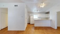 This single-story space showcases an open-concept layout that seamlessly connects the living area to the kitchen. The flooring throughout is warm hardwood-style planks, adding a natural, cohesive feel to the interior. The kitchen is outfitted with white cabinetry, a white refrigerator, and a peninsula with a light-toned countertop that serves as a functional divider between the cooking and living spaces. Track lighting above the kitchen provides targeted illumination, while a ceiling fan in the adjacent living area offers both light and air circulation. The walls and ceilings are painted white, contributing to a bright, airy atmosphere. A textured popcorn ceiling adds a traditional finish overhead. A louvered closet door is visible along the hallway, offering convenient storage access. A window near the kitchen allows natural light to filter into the space. HVAC vents are positioned along the walls, ensuring efficient climate control throughout. The overall design is clean and functional, with a neutral palette that provides a versatile backdrop.