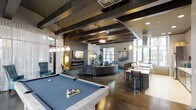 This single-floor amenity space is thoughtfully designed to blend social entertainment with relaxed comfort. Exposed dark wood ceiling beams add rustic character to the otherwise modern interior, while an array of pendant and track lighting creates a warm, inviting ambiance throughout. The open-plan layout is anchored by a full-size pool table with a blue felt surface, making it an immediate focal point upon entry. Adjacent to it, a generous circular lounge area features a curved sectional sofa set atop a teal area rug, centered around a sleek wall-mounted fireplace — ideal for gathering and conversation. A bar-height counter with seating provides a casual dining or socializing area, complemented by a well-appointed wet bar or kitchenette nearby. Large industrial-style windows along the perimeter allow abundant natural light to pour in, offering views of the surrounding outdoors. The flooring throughout is rich hardwood, adding warmth and continuity across the space. Curated artwork adorns the walls, and carefully chosen accent furniture in teal and gold tones contribute to the space's contemporary yet comfortable aesthetic. The overall design strikes a balance between a vibrant entertainment hub and a welcoming social retreat.