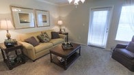 This single-story space showcases a thoughtfully designed living room that combines comfort and classic elegance. The room is anchored by a plush, beige sofa accented with patterned throw pillows, paired with a dark wood coffee table featuring a lower shelf for storage — a practical yet stylish centerpiece. Soft, neutral-toned carpeting extends throughout the space, contributing to a warm and inviting atmosphere.

Crown molding lines the ceiling, adding a refined architectural detail, while a chandelier overhead provides ambient lighting. A large window fitted with horizontal blinds allows ample natural light to fill the room, creating an airy feel. The window's placement suggests access to an outdoor area, such as a patio or balcony.

A dark wood end table with a turned-leg design holds a classic table lamp, offering warm accent lighting. Two gold-framed mirrors are displayed on the wall, enhancing the sense of depth and reflecting light throughout the room. The overall aesthetic is traditional and polished, with a neutral color palette of beige, taupe, and brown tones creating a cohesive and welcoming environment.