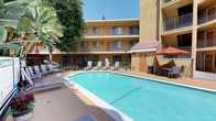This property is organized around a welcoming interior courtyard that serves as its central outdoor amenity space. The building's warm golden-yellow stucco exterior, accented by a red tile-roofed entryway feature and bold orange-red unit doors, gives the property a distinctive Southwest-inspired aesthetic. Multiple floors of units overlook the courtyard, each level accessible via open-air walkways lined with black metal railings and balconies that provide residents with direct views of the outdoor space below.

The centerpiece of the courtyard is a generously sized rectangular swimming pool with clear turquoise water, surrounded by a tan-colored concrete sun deck. Multiple lounge chairs are positioned poolside, offering ample space for sunbathing. A shaded outdoor dining area with a red market umbrella, table, and chairs provides a comfortable spot for al fresco relaxation. Mature trees and tropical plantings, including palm and banana-leaf varieties, are visible beyond the fence line, contributing greenery and shade. The courtyard is enclosed by a wrought-iron fence, creating a private and secure recreational environment. Overall, the space exudes a bright, resort-like ambiance well-suited to warm-climate living.