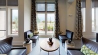 This single-floor community or amenity space is thoughtfully designed to accommodate a variety of uses, blending contemporary aesthetics with functional versatility. The layout is open and expansive, with distinct zones that flow seamlessly into one another, creating a cohesive yet dynamic environment.

Floor-to-ceiling windows flood the lounge areas with natural light and frame views of the surrounding landscaping and streetscape. The lounge zone features plush upholstered seating, a round wood coffee table, and geometric accent décor, all anchored beneath soaring window walls dressed with patterned drapes.

Multiple conference and co-working tables are positioned throughout the space, each surrounded by leather-upholstered chairs in a warm cognac tone. One dining-style conference area is highlighted by a shiplap accent wall with integrated wall sconces and a wall-mounted flat-screen display, creating a polished and professional atmosphere.

Structural columns punctuate the open floor plan, while sliding barn-style doors offer flexible room division. Accent walls in muted olive and deep charcoal introduce pops of color, complemented by exposed brick-like stone detailing near the entrance. The light-toned carpeting and neutral ceiling palette tie the entire space together, resulting in a warm, inviting, and well-appointed environment suited for both collaboration and relaxation.
