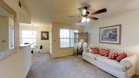 This single-floor residence offers a welcoming and well-appointed interior designed for comfortable everyday living. The main living area is a standout space, featuring vaulted ceilings that lend a sense of openness and airiness. A wood-blade ceiling fan with integrated lighting is centrally mounted, providing both functionality and a classic aesthetic touch. The room is finished with plush wall-to-wall carpeting in a neutral tone, complementing the warm beige walls that carry throughout the space. A window fitted with horizontal blinds and curtain panels allows natural light to filter in, brightening the room while maintaining privacy. Adjacent to the living area, a dining nook is visible, furnished with a small bistro-style table and chairs beneath a decorative pendant light, making it an intimate spot for meals. A framed mirror and framed artwork add decorative character to the walls. The layout flows naturally between the living and dining spaces, creating a cohesive and connected feel. The overall ambiance is cozy and inviting, with warm lighting and classic décor details that contribute to a comfortable, lived-in atmosphere throughout the home.