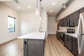 This contemporary two-story space offers a thoughtfully designed layout with modern finishes throughout. The open-concept kitchen serves as the heart of the main level, anchored by a substantial island with a white quartz countertop, an integrated sink, and a slate-blue base. Dark wood shaker cabinetry lines the walls, complemented by a classic white subway tile backsplash and a full suite of stainless steel appliances. Exposed industrial-style ductwork adds an urban character to the space, while pendant lighting with clear glass shades provides warm, focused illumination over the island. Light hardwood flooring runs seamlessly throughout both levels, enhancing the bright and airy atmosphere created by the neutral white walls. The bedrooms are generously sized, each featuring a ceiling fan and walk-in or reach-in closets with built-in shelving. One bedroom offers direct access to a private balcony with outdoor views. The bathrooms are finished with dark wood vanities, white countertops, and modern fixtures. A staircase with black metal balusters connects the two floors, contributing to the space's clean, contemporary aesthetic. Recessed lighting and black-framed windows complete the refined, modern look found throughout.